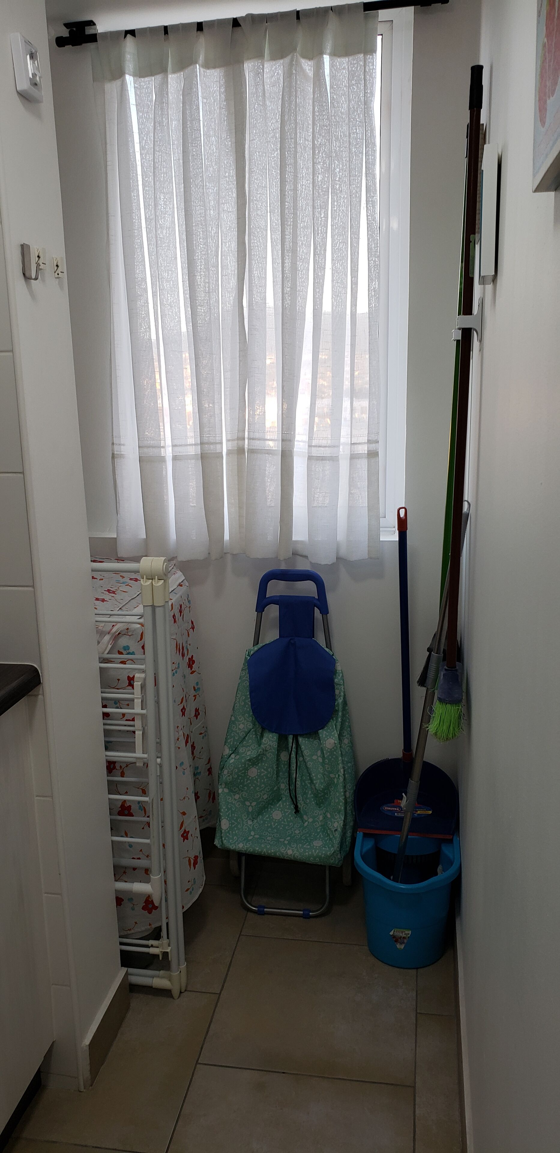 Una habitación pequeña con una ventana cubierta por cortinas traslúcidas, un radiador con funda floral, un cubo de limpieza azul y un trapeador apoyado contra la pared.