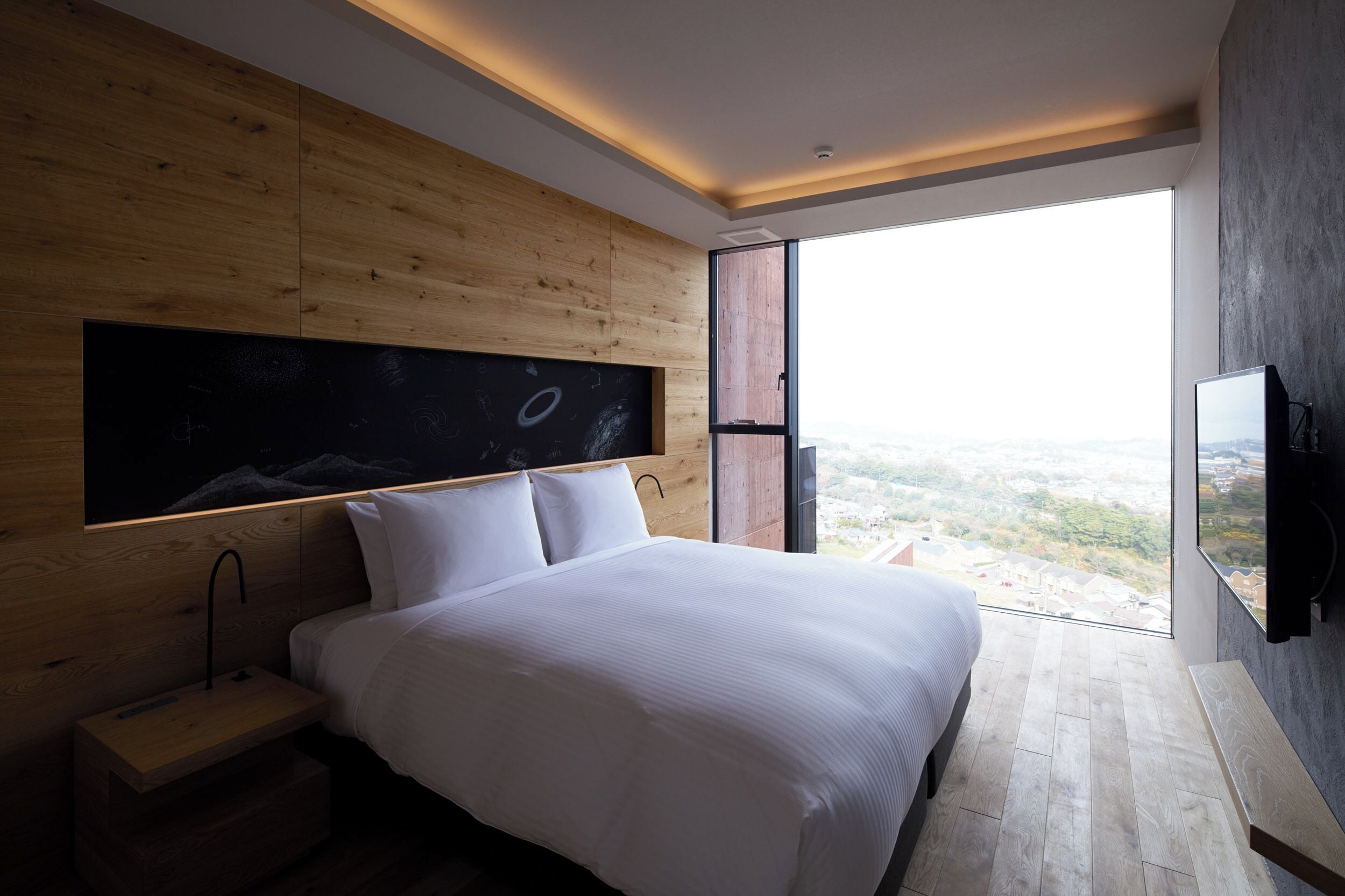 A modern bedroom with a large bed, wooden panel walls, a blackboard, and a city view.