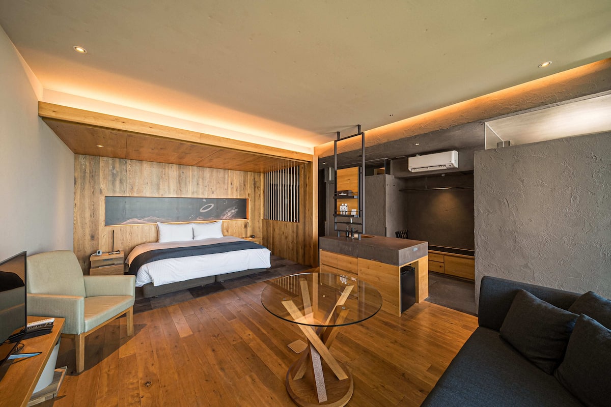 A modern hotel room with a wooden floor, a glass coffee table, a bed with a wooden headboard, a sofa, a desk with a chair, and a built-in wardrobe.