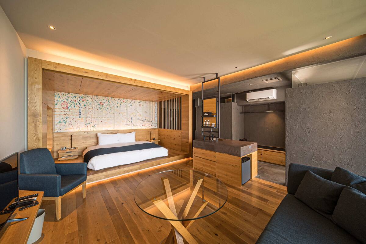 A modern hotel room with a wooden floor, a glass table, a bed with white linens, a blue armchair, a sofa, a map on the wall, and a kitchenette.