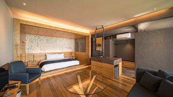 A modern hotel room with a wooden floor, a glass table, a bed with white linens, a blue armchair, a sofa, a map on the wall, and a kitchenette.