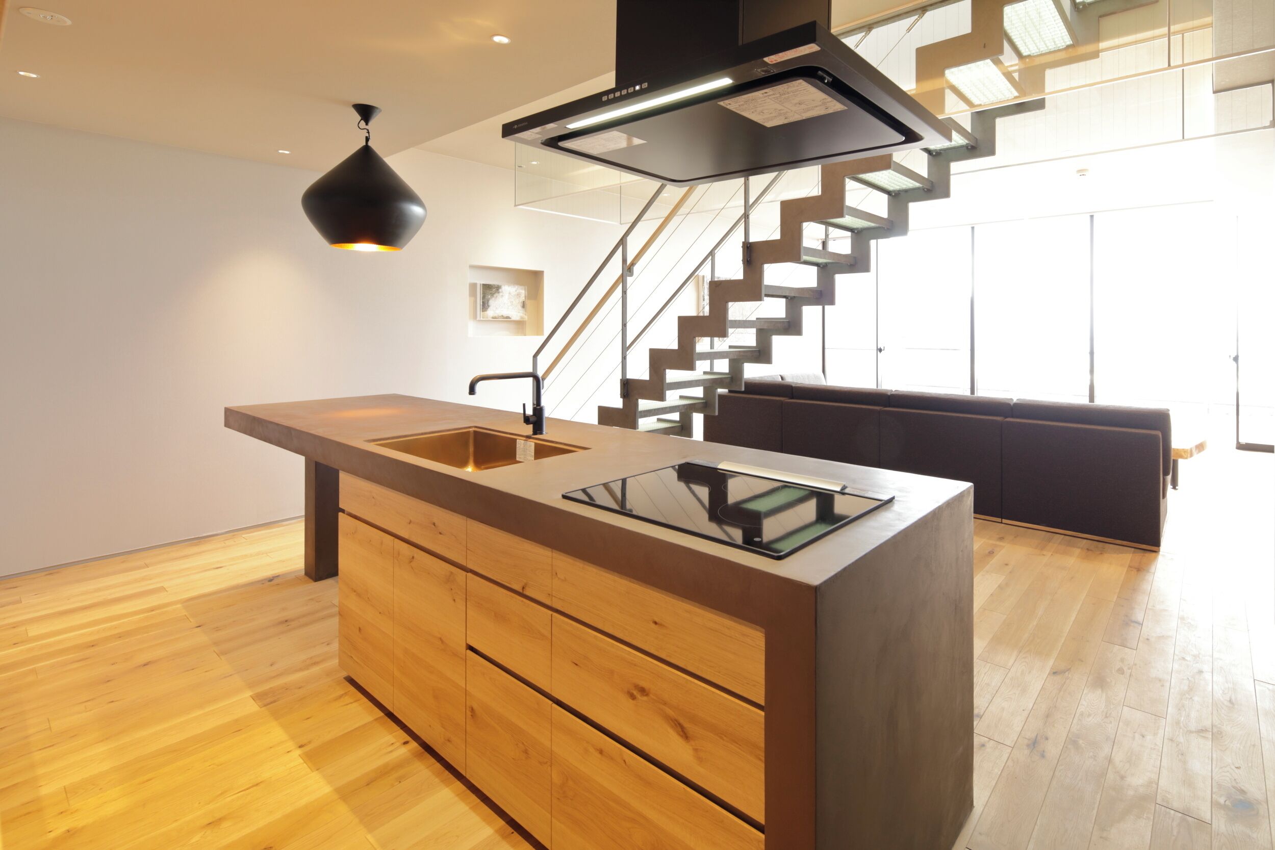 Una cocina moderna con una isla central, armarios de madera, una lámpara colgante negra y una escalera que sube a un nivel superior.