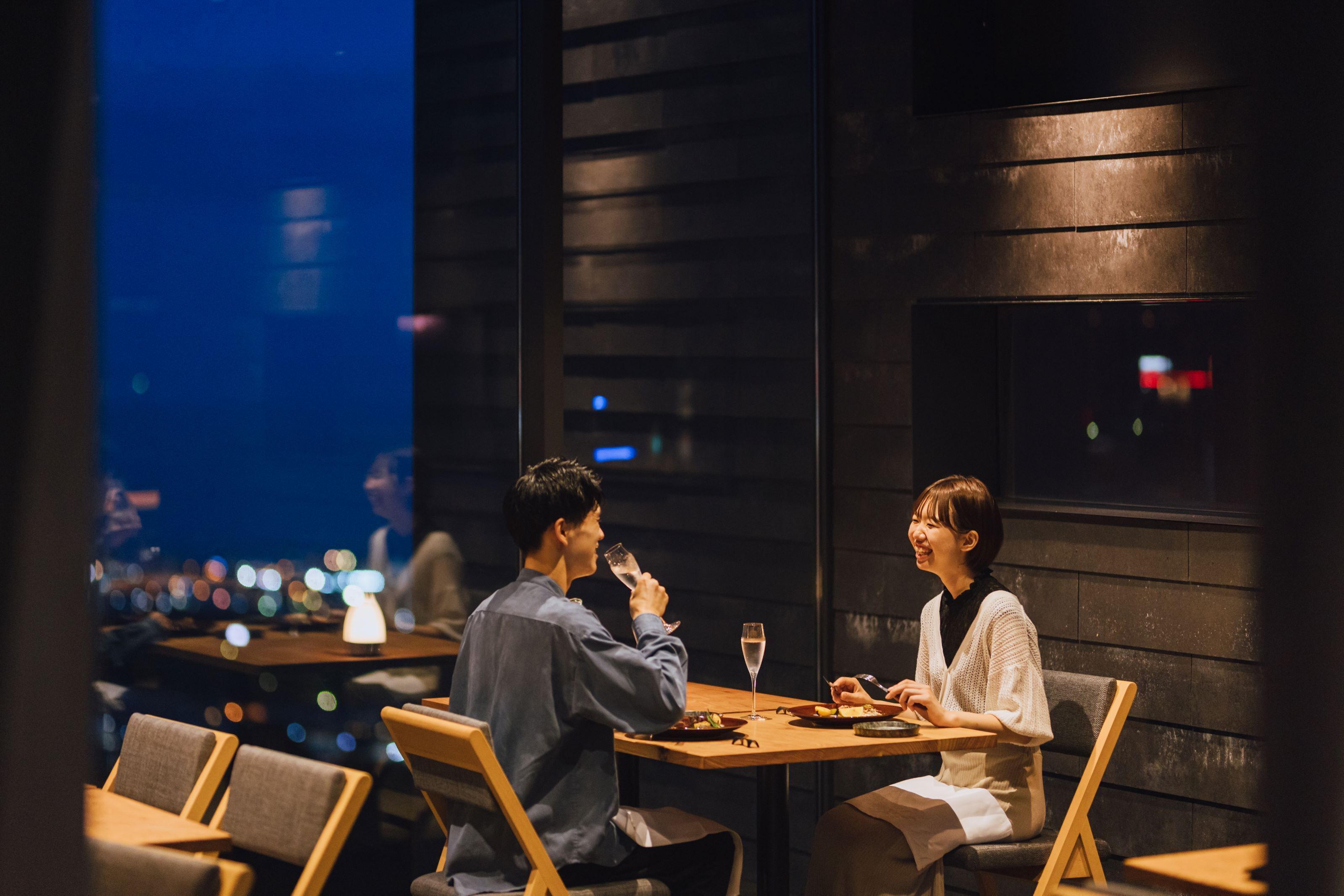 A couple dining at a table with a city view, wooden furniture, and dim ambient lighting.