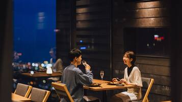 A couple dining at a table with a city view, wooden furniture, and dim ambient lighting.