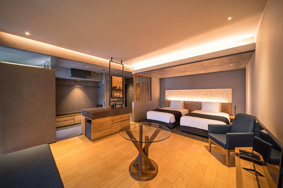 A modern hotel room with a glass-top table, two beds, a sofa, and a TV.