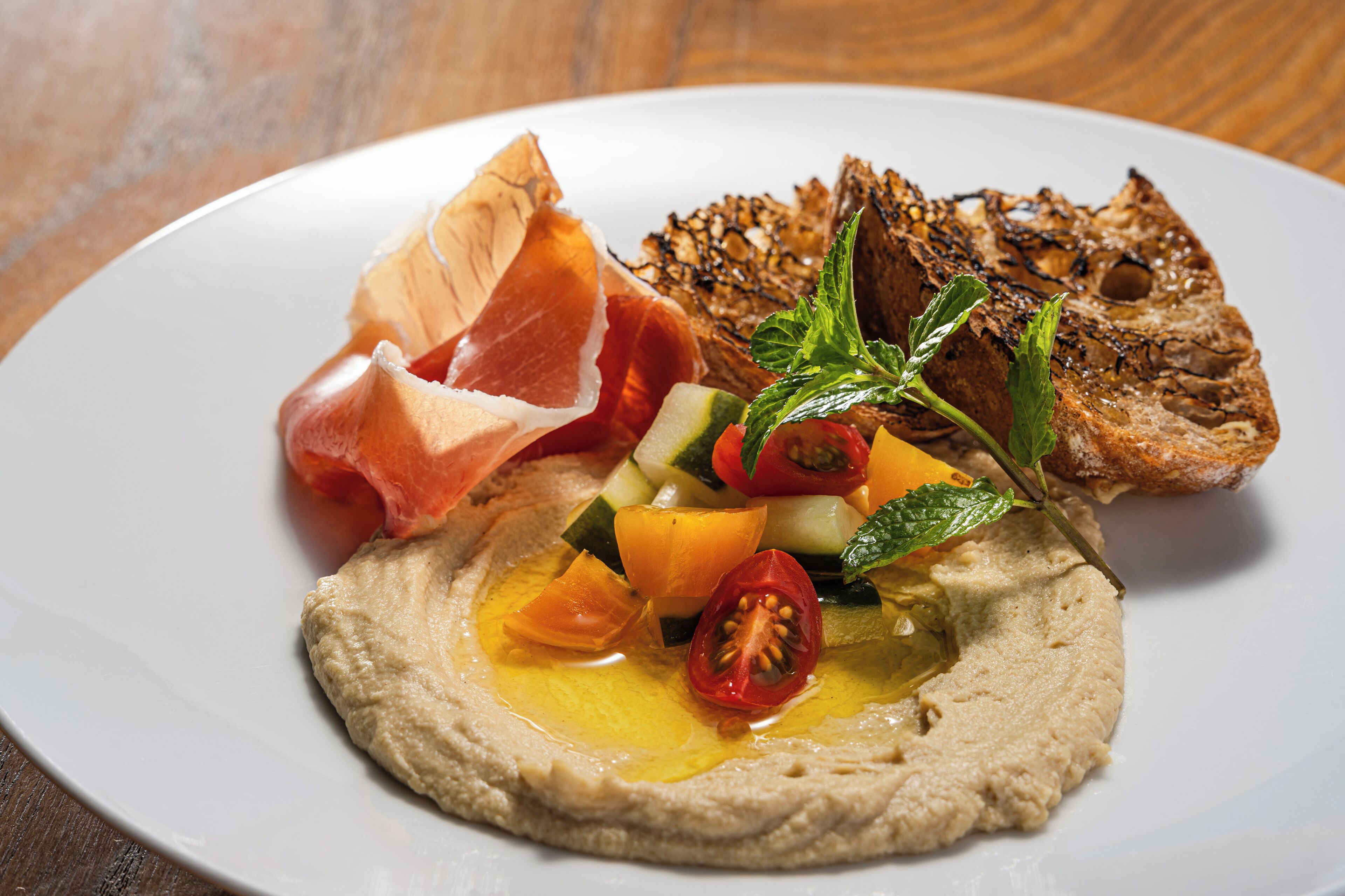 Een bord met hummus, gegrilde groenten, parmaham en geroosterd brood.