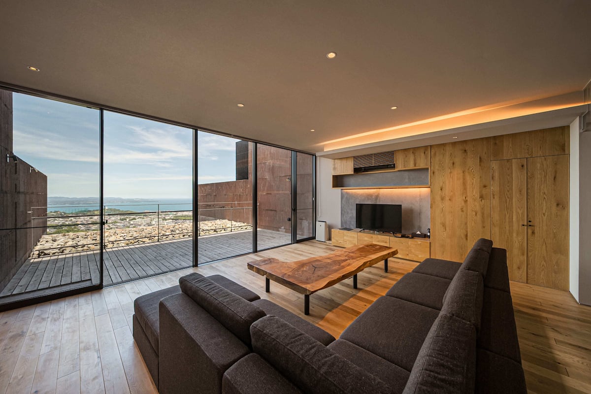A modern living room with a large wooden coffee table, dark sofas, and a television. A wooden cabinet and a fireplace are visible in the background.