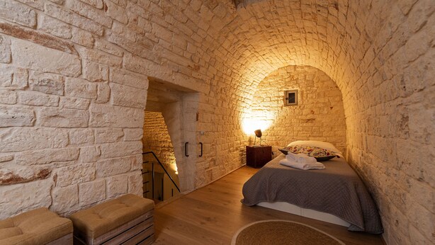 A cozy room with a stone archway, a bed with a quilt, a nightstand with a lamp, and a bench. - Trulli in corte (Alberobello)