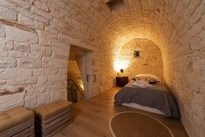 A cozy room with a stone archway, a bed with a quilt, a nightstand with a lamp, and a bench. - Trulli in corte (Alberobello)