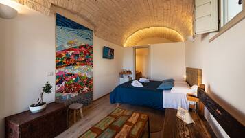 Un dormitorio con una cama, un banco, una mesita pequeña, una planta en maceta y un mural colorido en la pared.