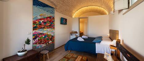 A bedroom with a bed, a bench, a small table, a potted plant, and a colorful mural on the wall.