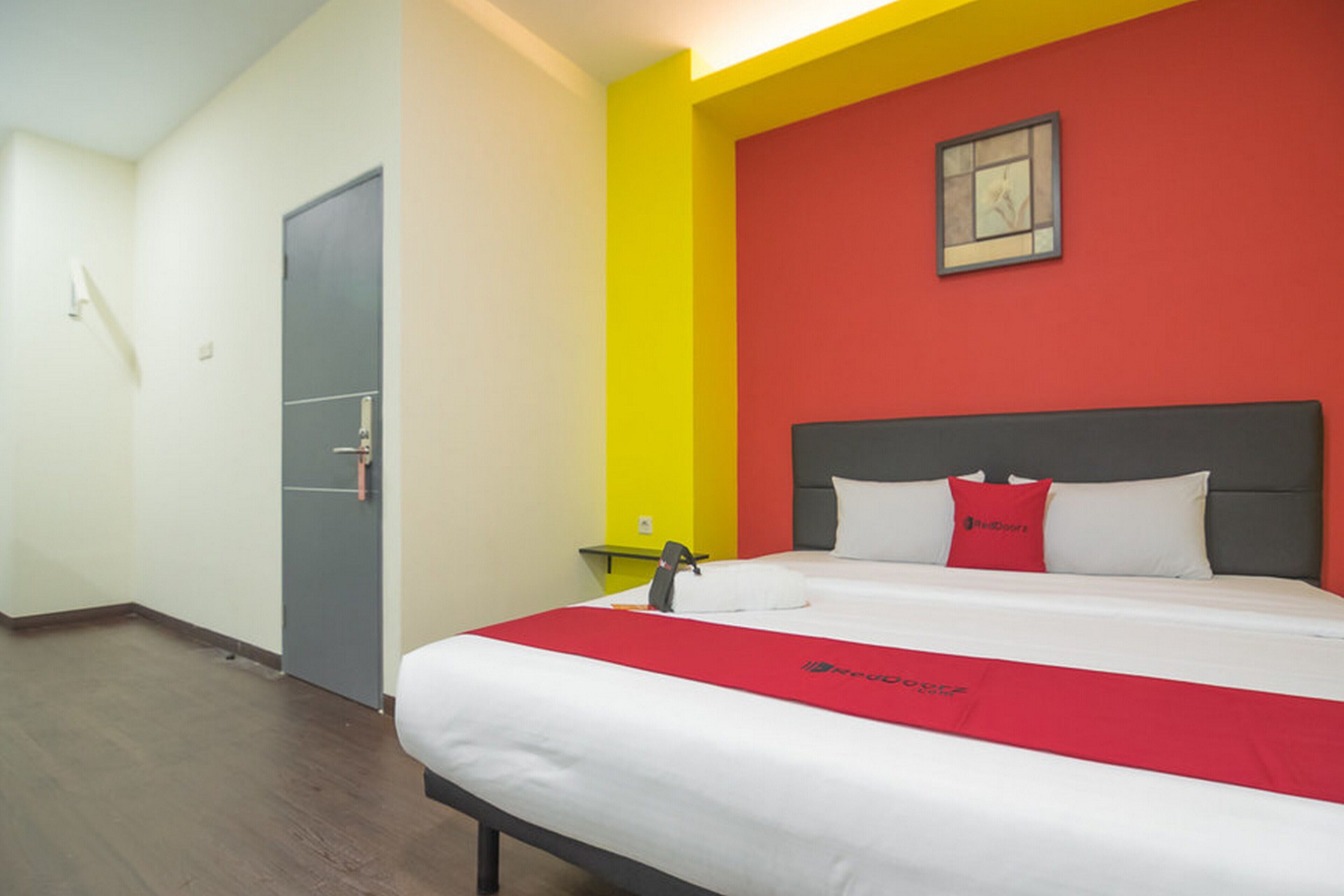 Une chambre d’hôtel avec un lit, une couvre-lit rouge et blanche, une porte grise et un mur rouge orné d’un tableau.