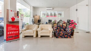 Un hall d’hôtel avec deux canapés beiges, un fauteuil à motifs floraux, un panneau RedDoorz, un bureau avec un ordinateur et une fenêtre ornée d’affiches.