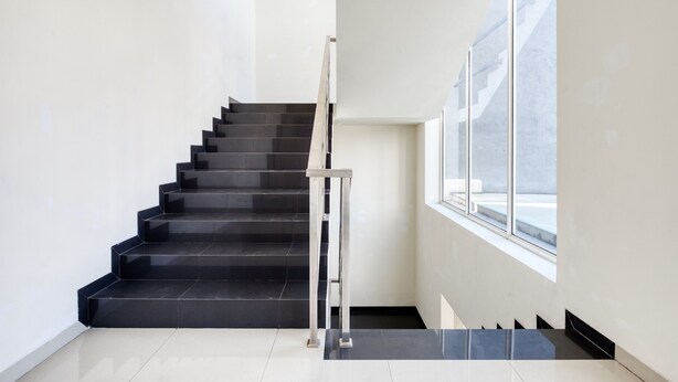 Un escalier avec des marches noires et une rampe métallique, menant à un palier doté d’une grande fenêtre.