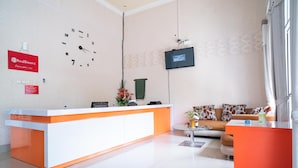 A reception area with a white and orange desk, a clock, a flat-screen TV, and a sofa with patterned cushions. - RedDoorz @ Jalan Pendidikan Mataram (Mataram)