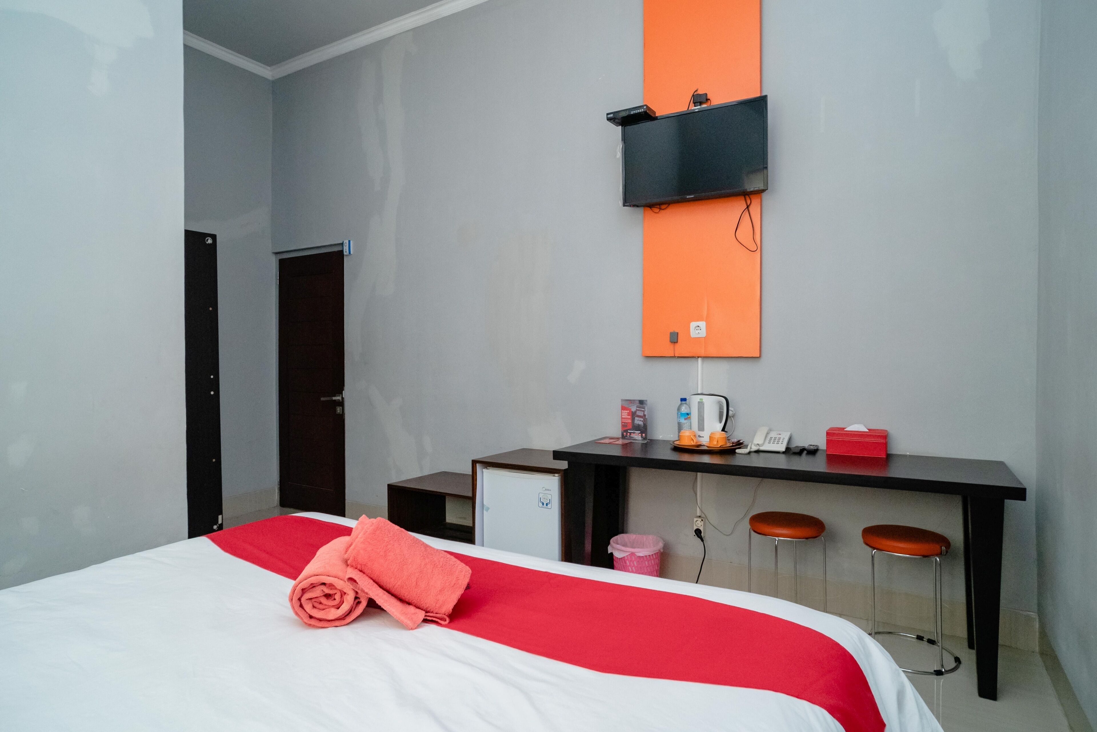 A hotel room with a bed, a desk with two stools, a small refrigerator, and a wall-mounted shelf.