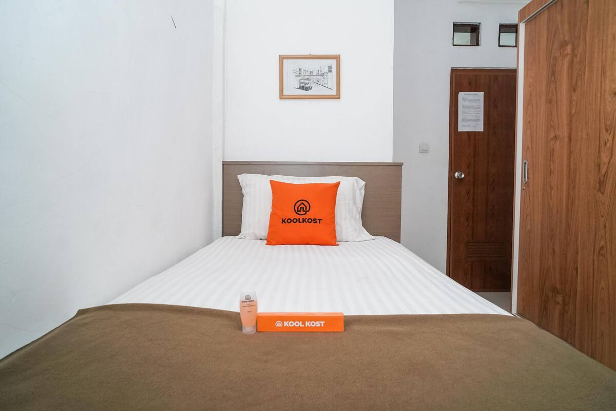 A neatly made bed with a white and orange pillow, a brown blanket, and a small box labeled "KOOLKOST" on the bed.
