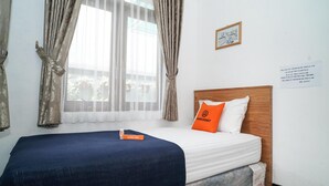 A neatly made bed with a blue bedspread and an orange pillow, a wooden headboard, and a window with sheer curtains. - KoolKost @ Batutulis Bogor (Bogor)
