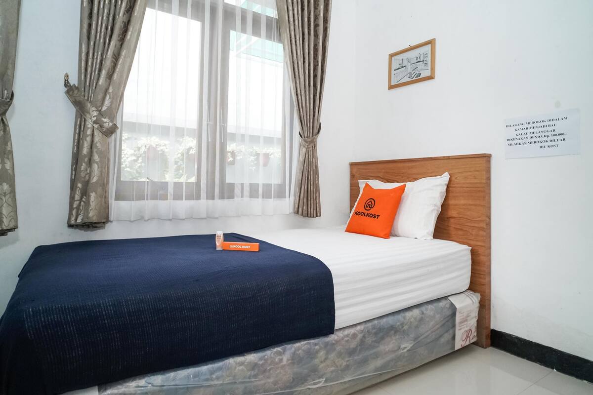 A neatly made bed with a blue bedspread and an orange pillow, a wooden headboard, and a window with sheer curtains.