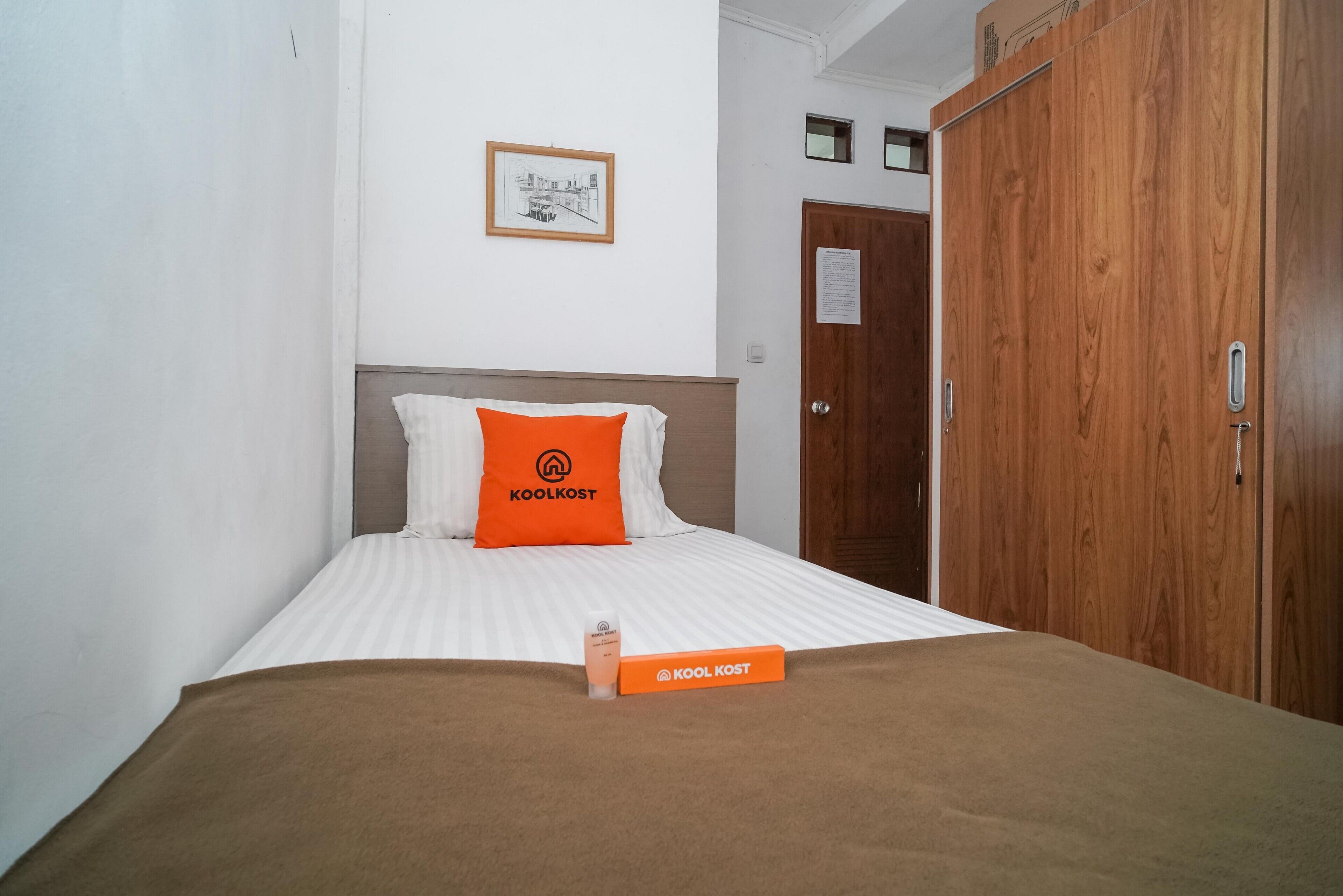 A hotel room with a bed, a brown blanket, a pillow with the brand 'Koolkost', and a wooden wardrobe.