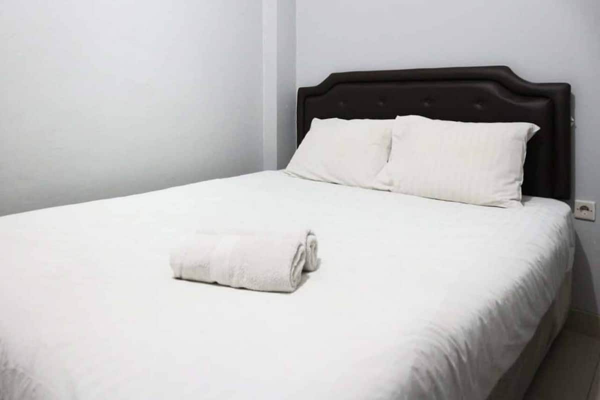 A neatly made bed with white linens and pillows, a rolled white towel on the bed, and a dark headboard.