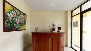 Un comptoir d’accueil en bois, équipé d’un ventilateur et de bouteilles d’eau, un tableau représentant des fleurs accroché au mur et une grande fenêtre.