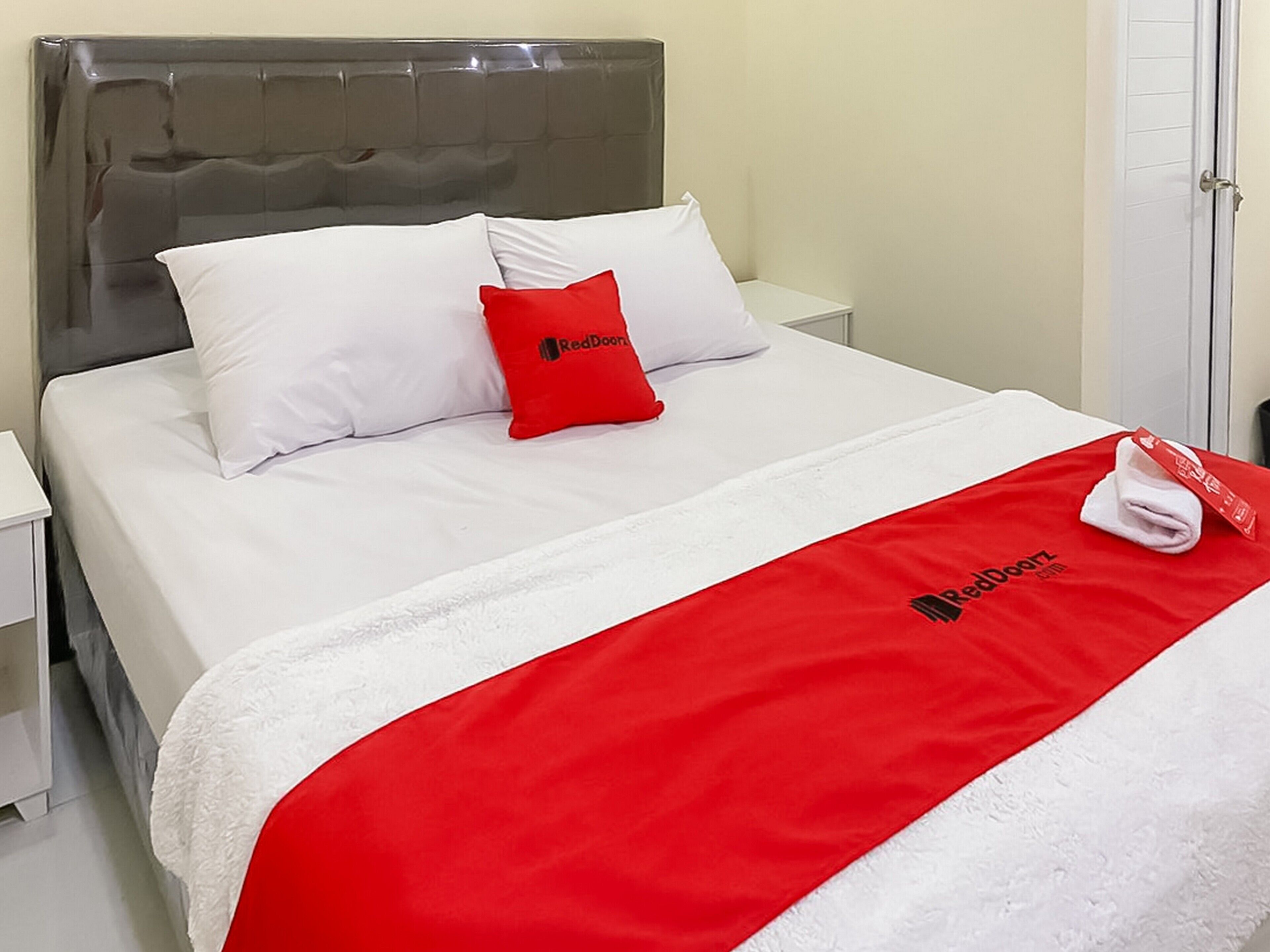 A neatly made bed with white bedding and a red blanket, a grey upholstered headboard, and a white nightstand with a lamp.