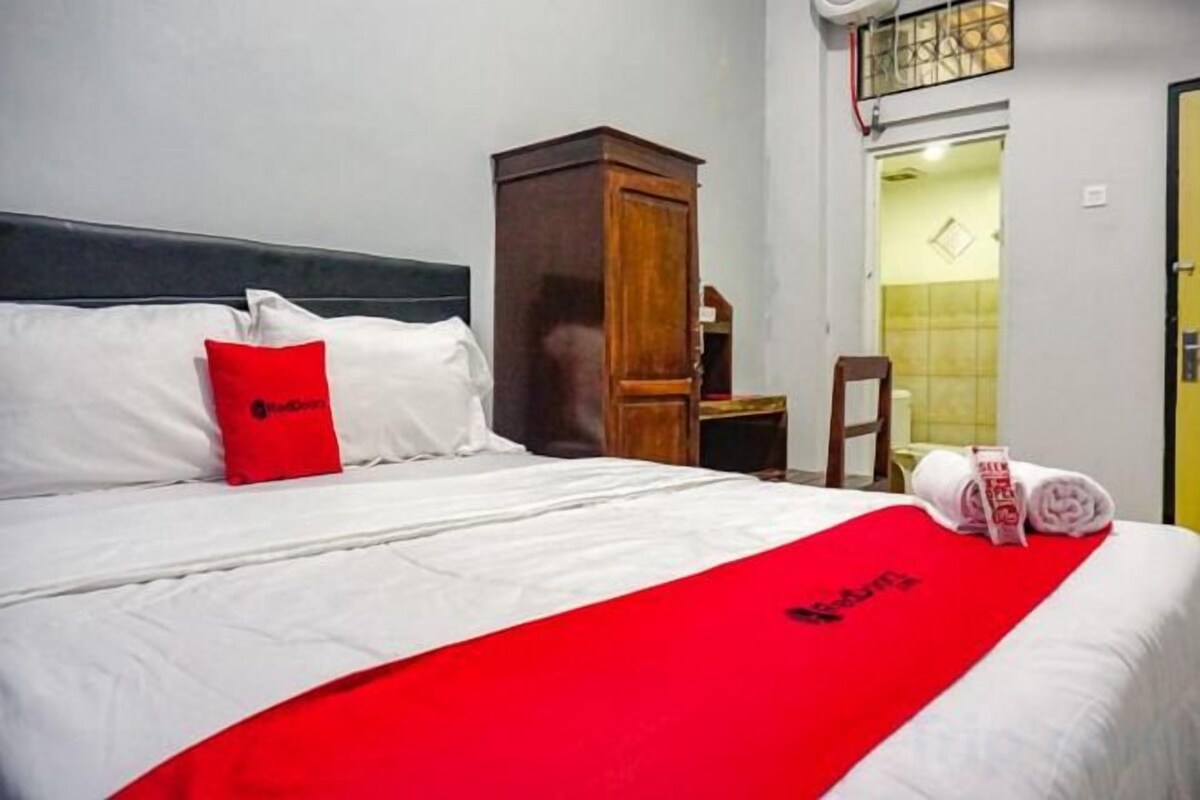 A hotel room with a bed, a wooden wardrobe, a red pillow, and a red blanket.