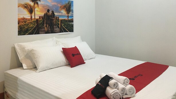A neatly made bed with white linens, a red pillow, and a red blanket with the RedBoutz logo. - RedDoorz near Institut Kesehatan Helvetia (Medan)