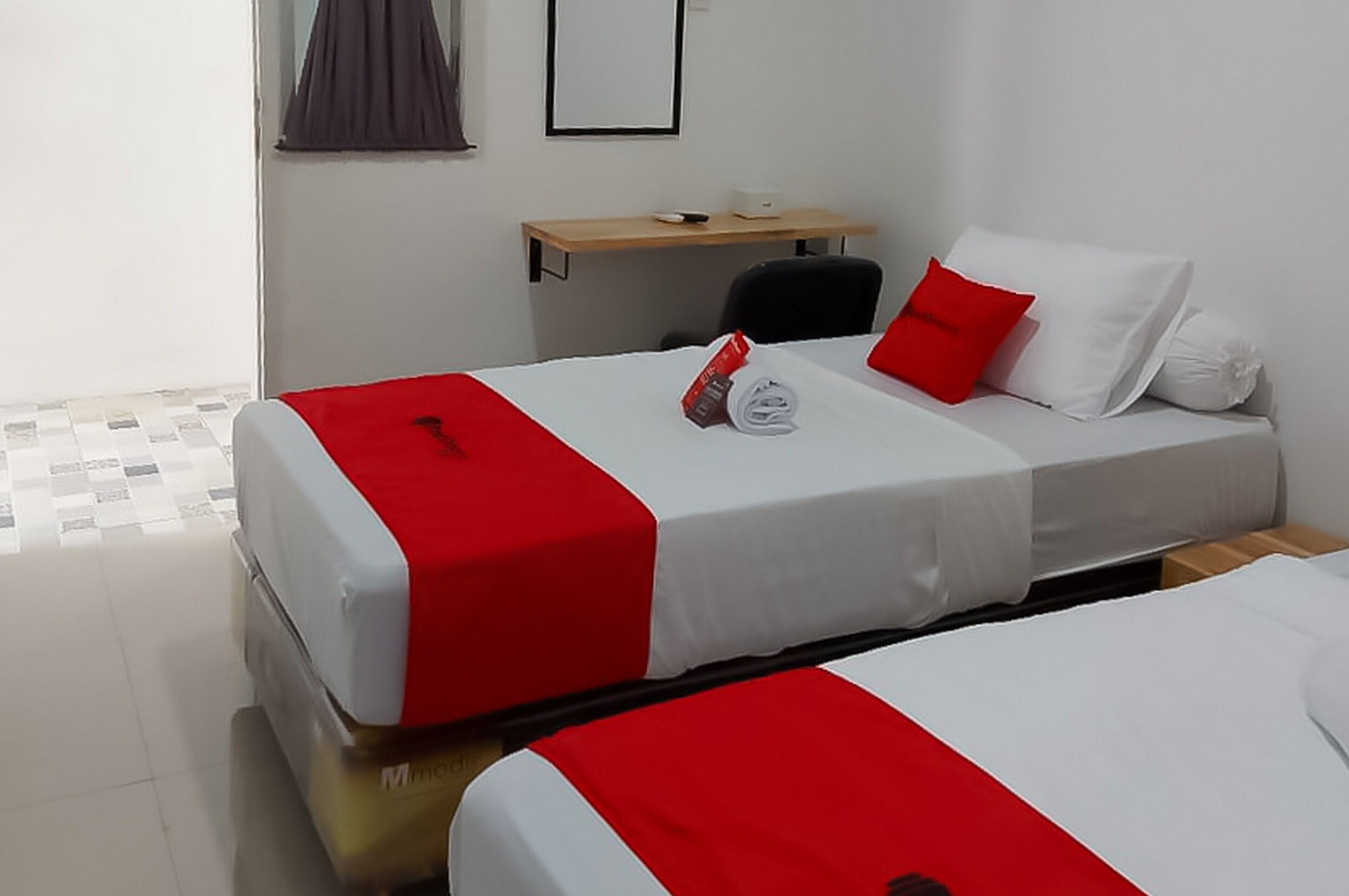 Hotelkamer met twee bedden, elk met rode en witte beddengoed, een bureau met stoel, en een klein tafeltje met een lamp.