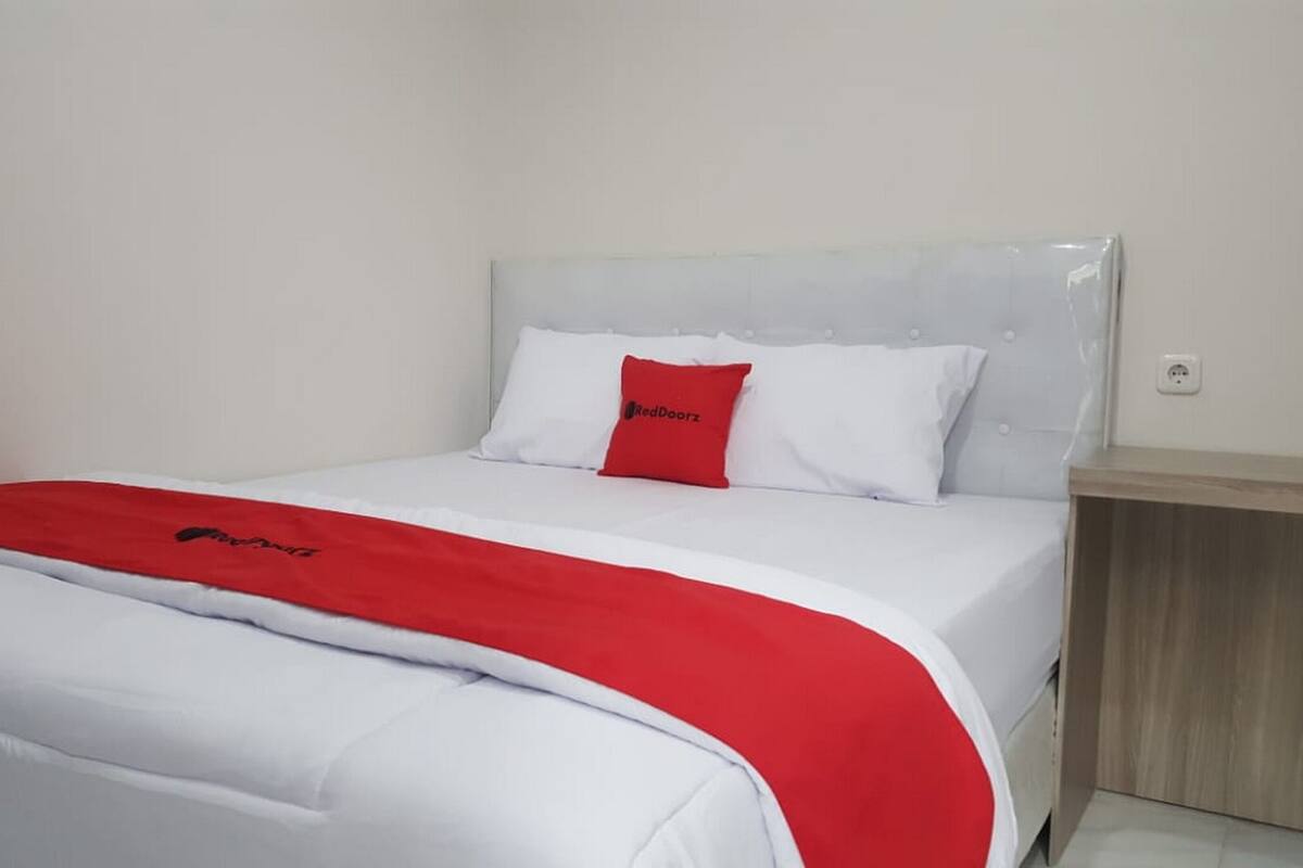Una cama con una colcha blanca y roja, un almohadón rojo y una mesita de noche de madera.