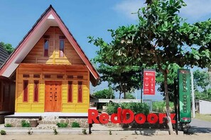 A wooden building with a red roof and yellow accents, a sign reading "RedDoorz", and a green sign with text.