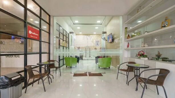 A modern hotel lobby with glass doors, a RedBook sign, and a RedBook logo. There are green and brown chairs around tables, and a trash bin is visible. - RedDoorz Plus @ Cemara Asri Medan (Medan)