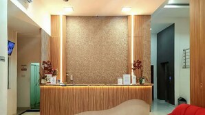 A reception desk with a wooden finish and a textured wall behind it. There is a red chair to the right of the desk. - RedDoorz Plus @ Cemara Asri Medan (Medan)