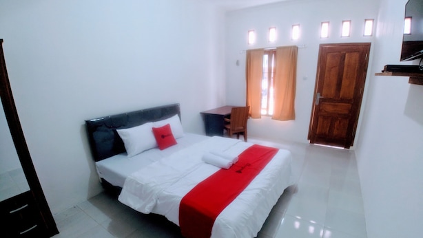A neatly made bed with white and red bedding, a wooden headboard, and a small desk with a chair. - RedDoorz near Exit Toll Jatiwarna 2 (Bekasi)