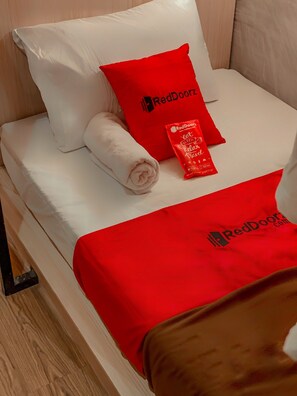 A neatly made bed with white linens, a red pillow with the RedDoorz logo, and a red blanket with the same logo.