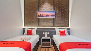 A hotel room with two beds, each with a red and white bedspread, a small bedside table with a lamp, and a picture of a sunset over a bridge on the wall. - RedDoorz Plus @ Doorman Guest House (Bandung)