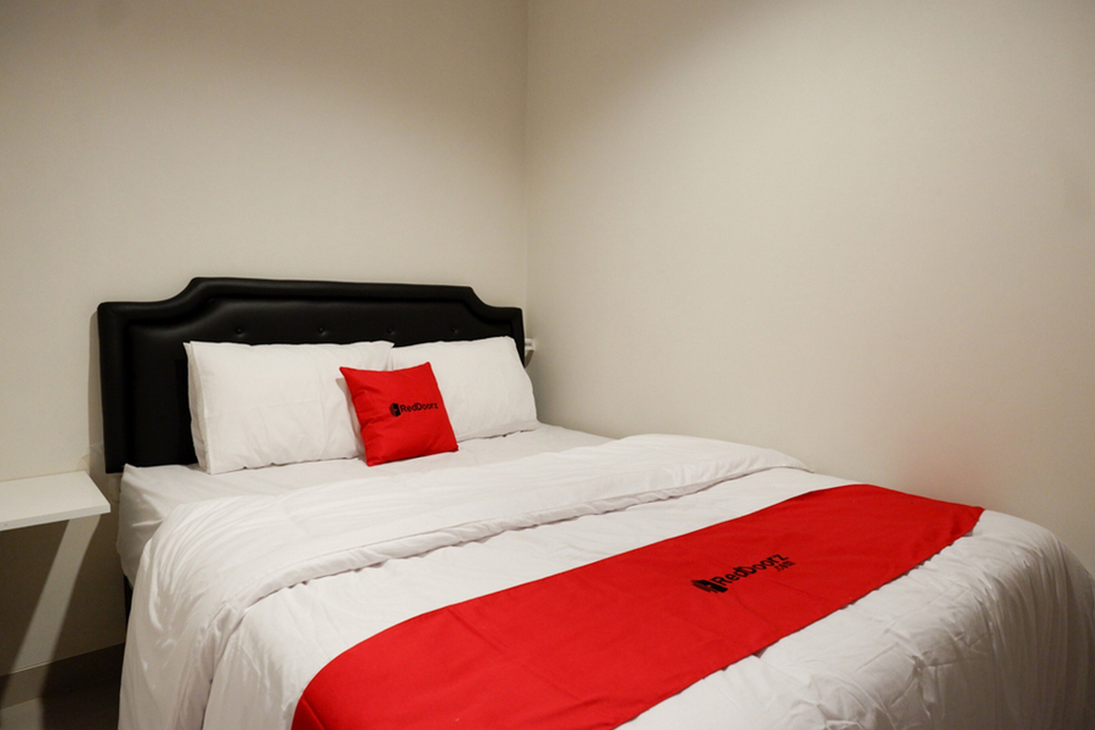 Una cama con ropa de cama blanca, un cojín de acento rojo y una manta roja con un logotipo.