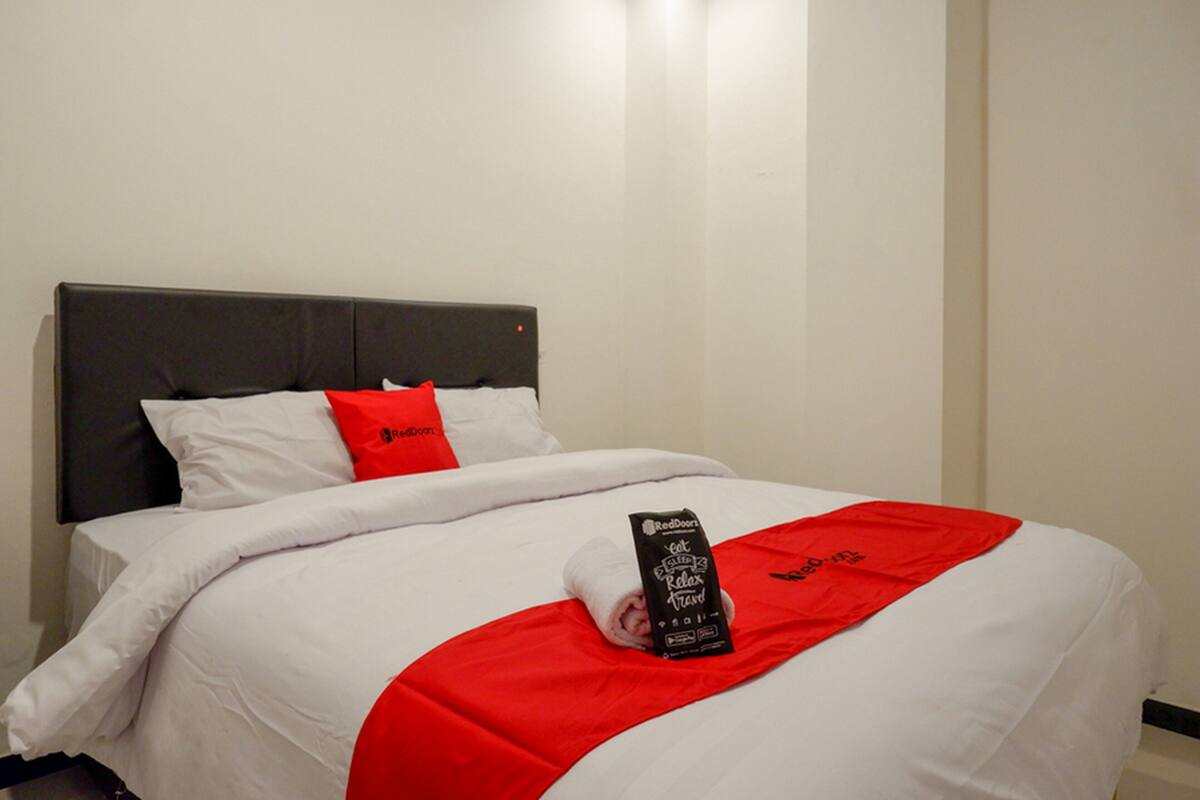 A neatly made bed with white and red bedding, a grey headboard, and a red towel with text on it.