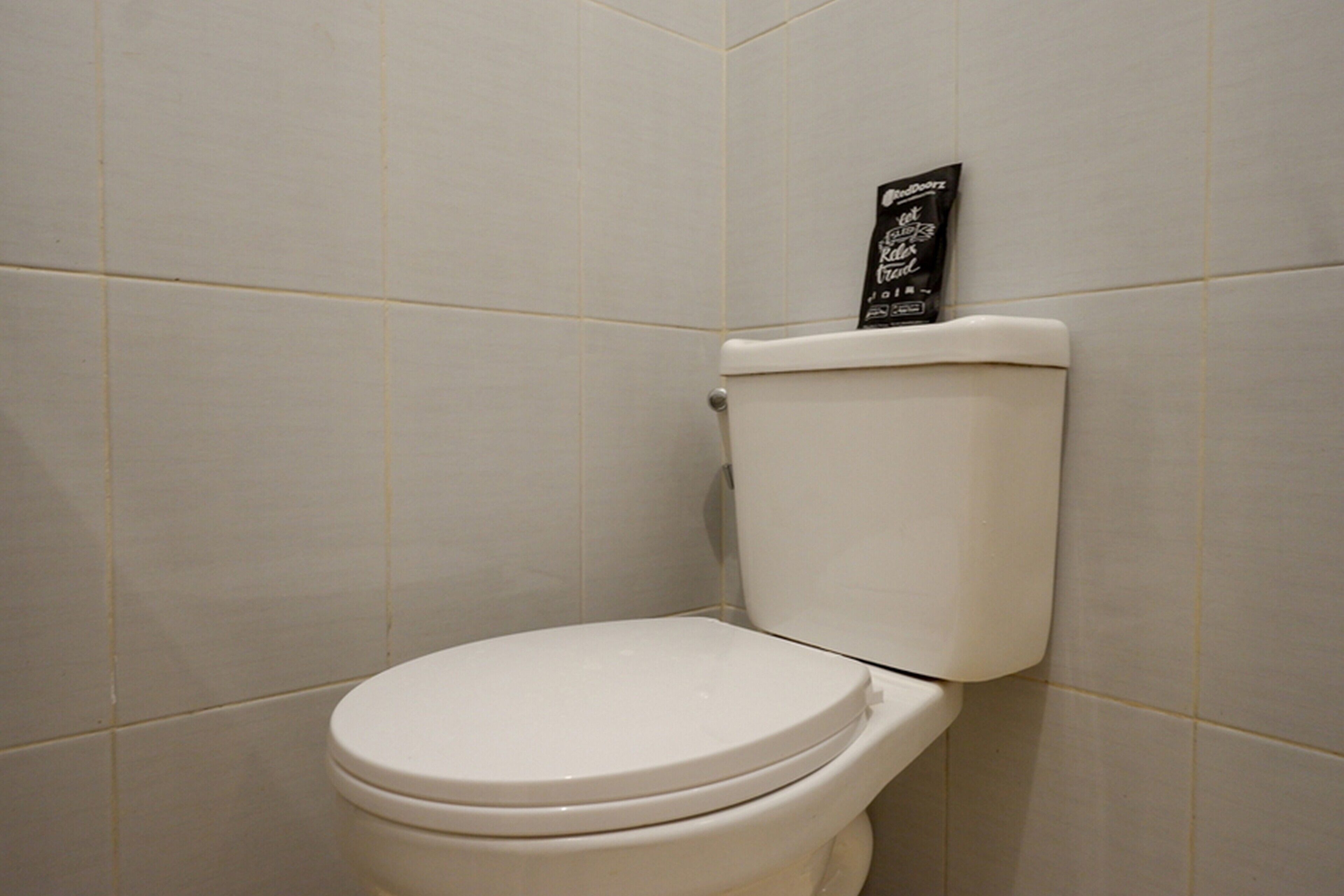 Een toilet met een witte deksel en tank, een zwart bordje op de tank, en witte betegelde wanden.