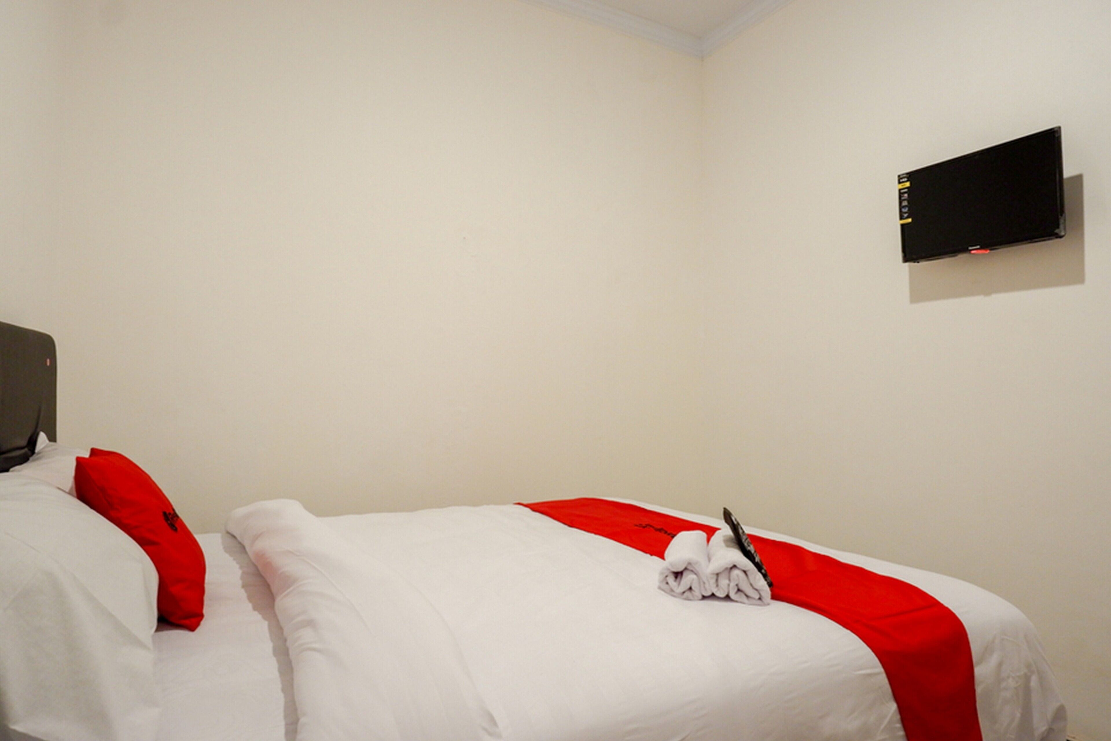 A hotel room with two beds, each with white bedding and red accents, a flat-screen TV mounted on the wall, and a neatly folded towel on the bed.