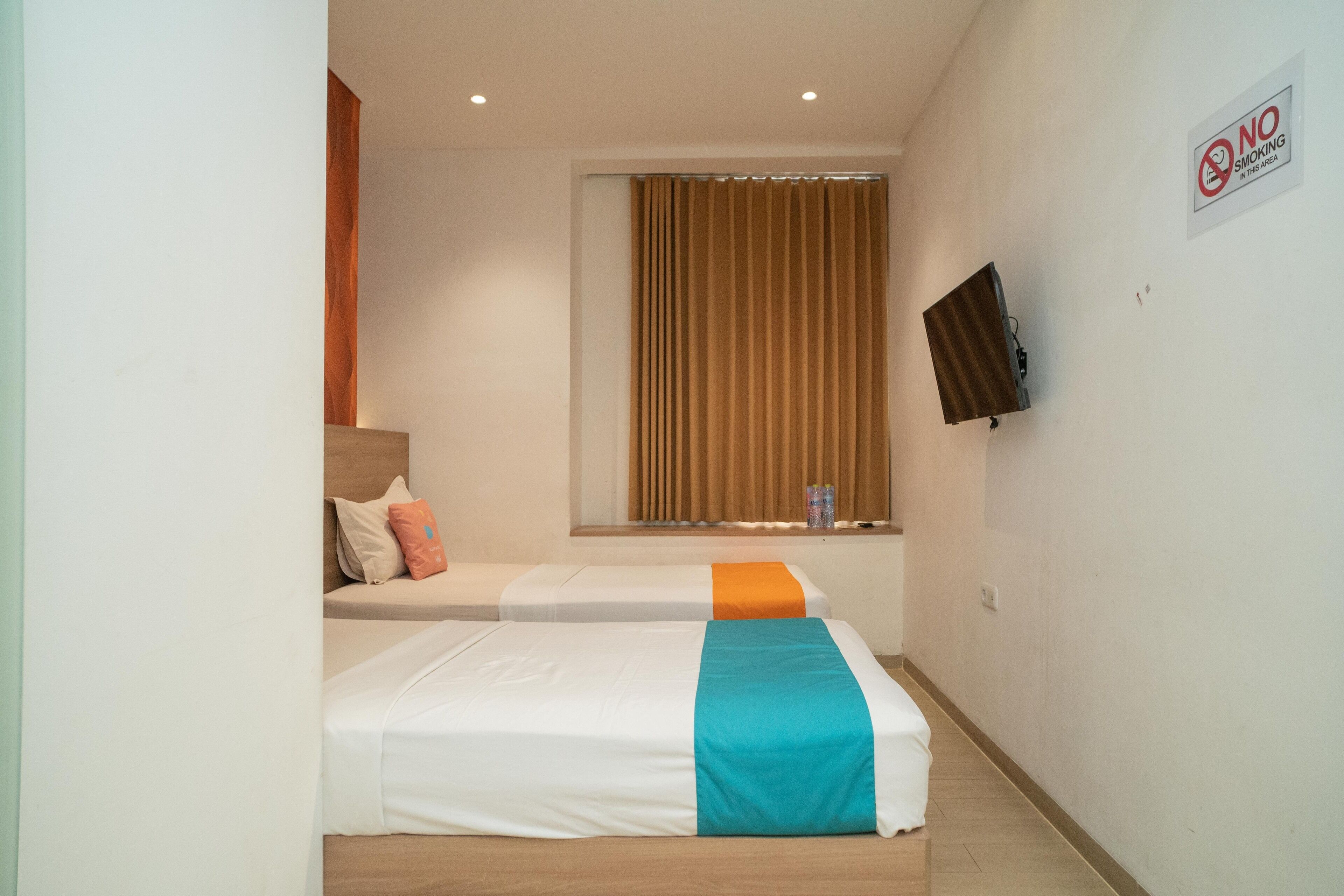 Une chambre d’hôtel avec deux lits, un téléviseur fixé au mur, une fenêtre avec des rideaux et un panneau indiquant « Il est interdit de fumer ».