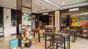 A modern restaurant with a clean, minimalist interior, featuring wooden tables and chairs, a decorative flower arrangement, and a visible menu board. - Sans Hotel Kupang Indah Surabaya (Surabaya)