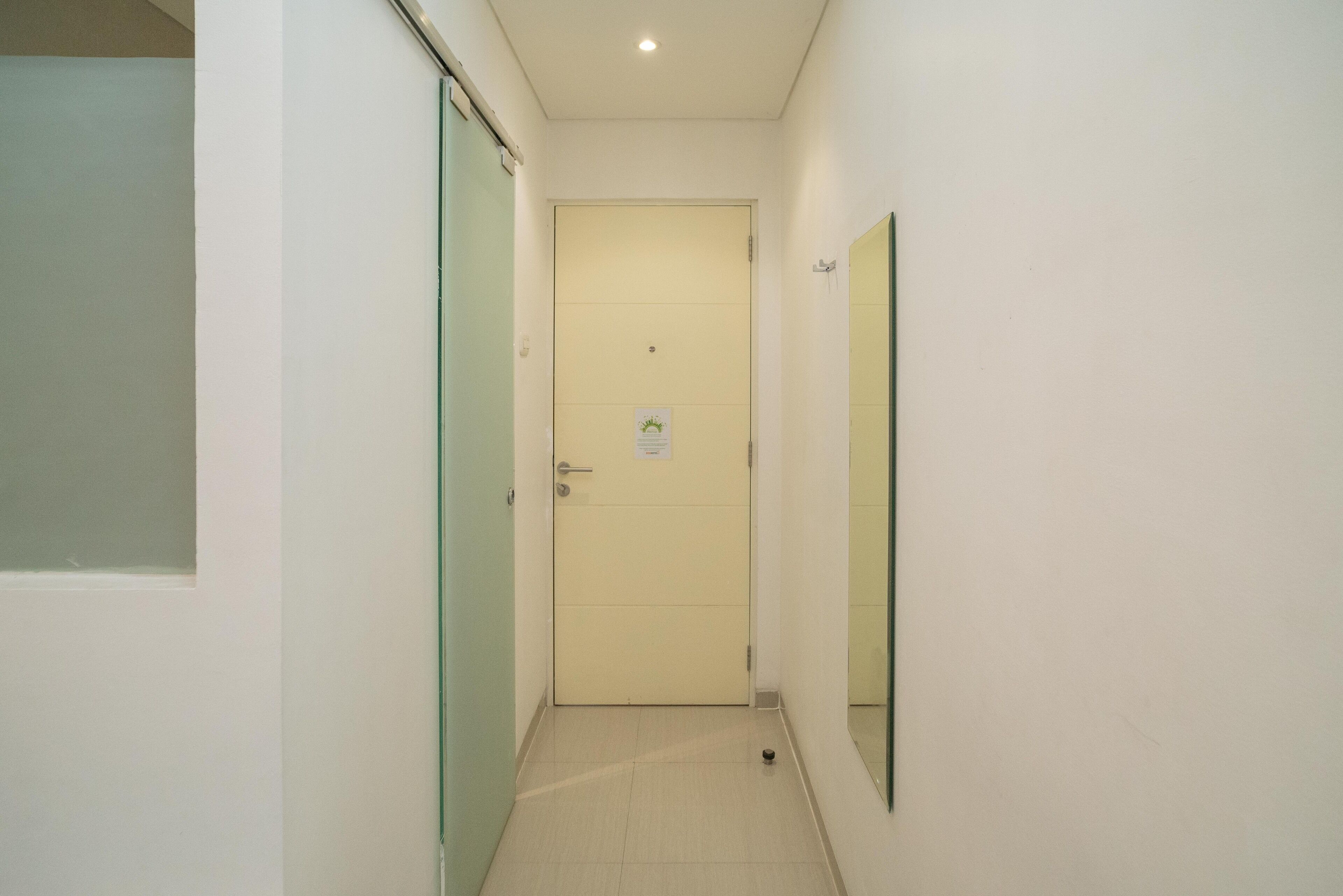Un pasillo estrecho con una puerta cerrada al final, una pequeña señal en la puerta y un espejo en la pared derecha.