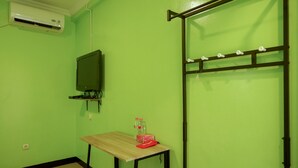 Ein kleines Zimmer mit grüner Wand, einem Holztisch, einem Hocker, einem an der Wand befestigten Fernseher und einem Wäscheständer.