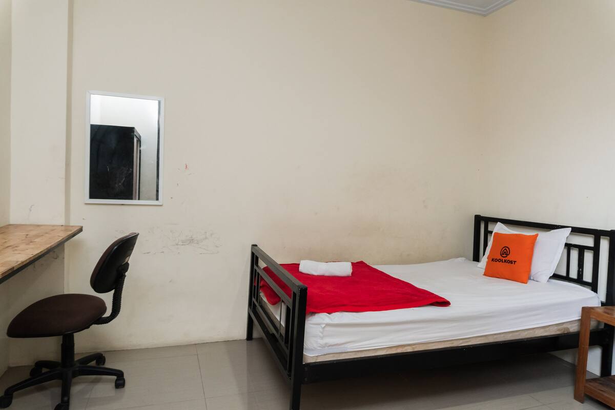 Een kleine kamer met een enkel bed, een houten bureau, een stoel en een spiegel aan de muur.