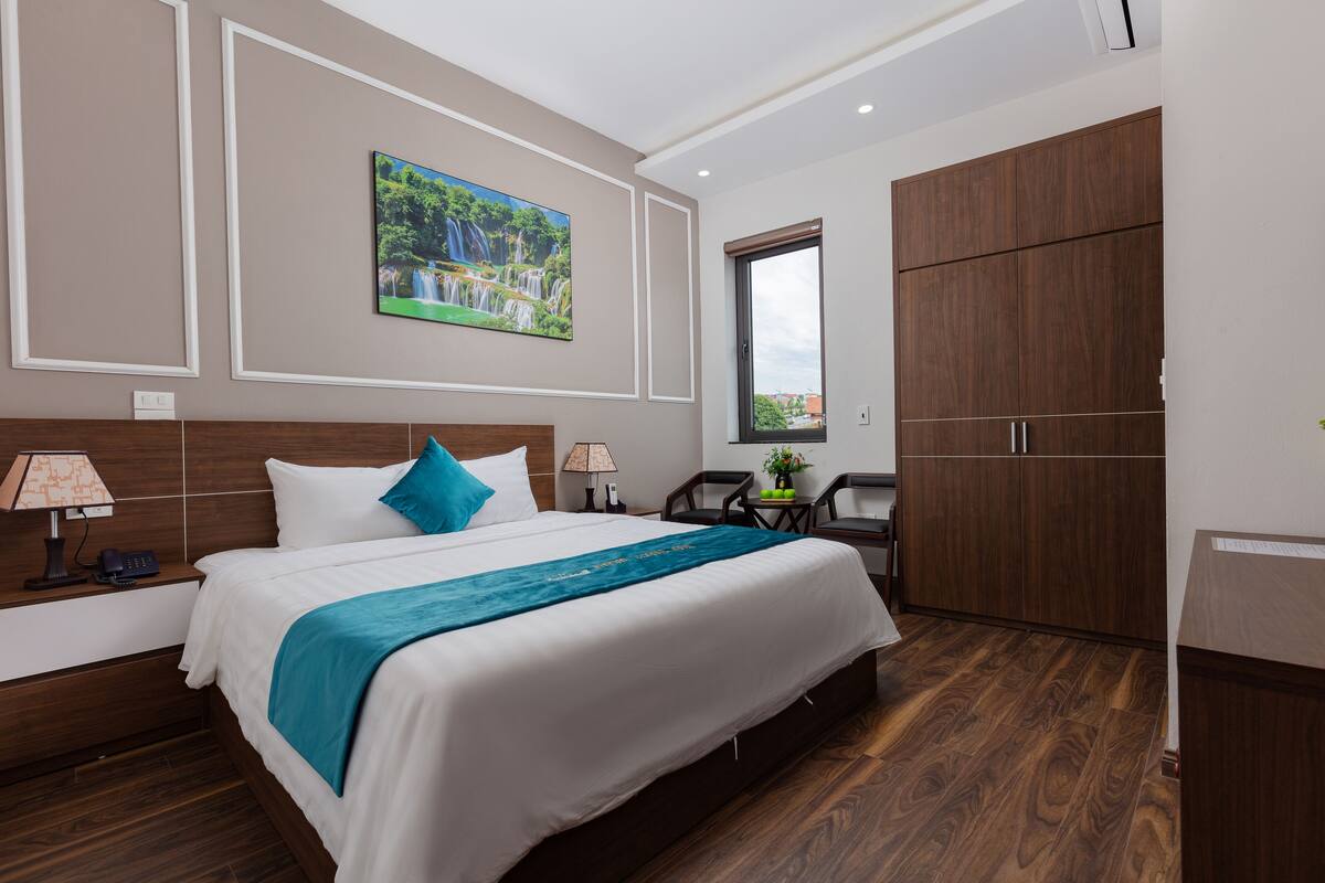 A hotel room with a large bed, a painting of a waterfall, wooden furniture, and a window with a view.
