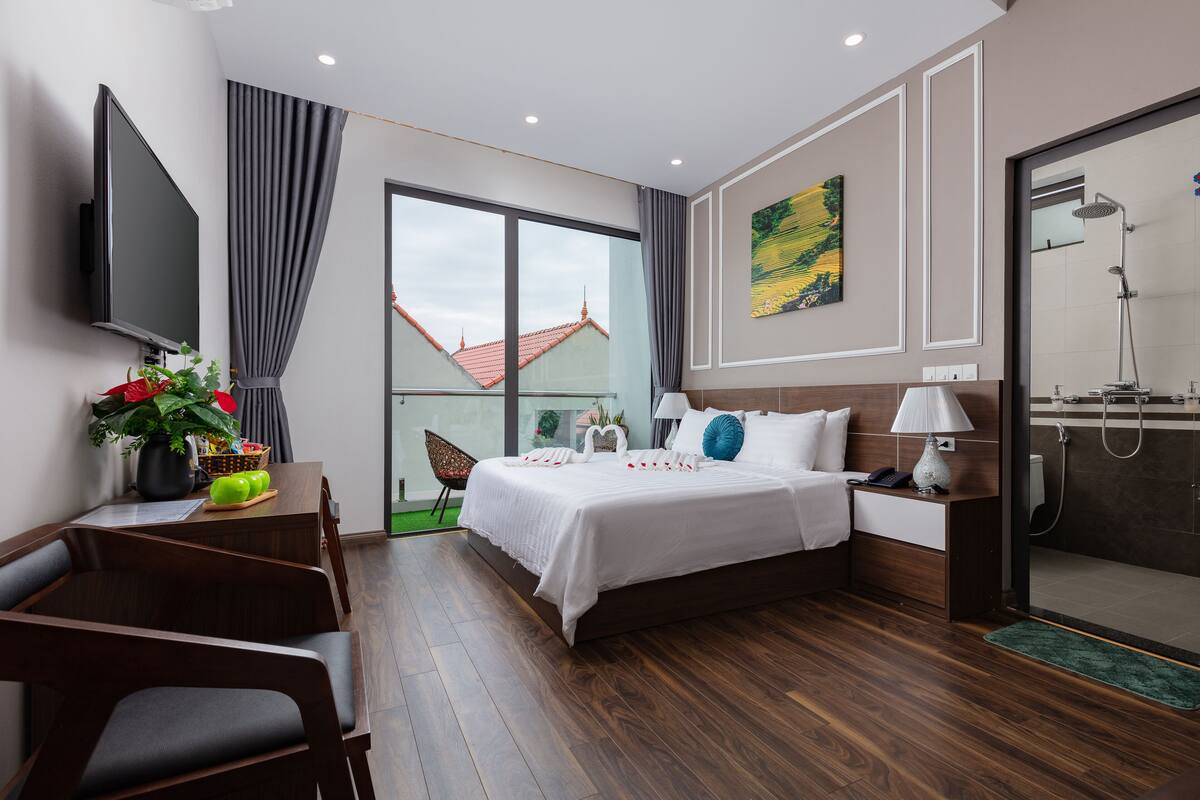 A modern hotel room with a large bed, a desk with a flat-screen TV, a shower area, and a view of buildings outside.