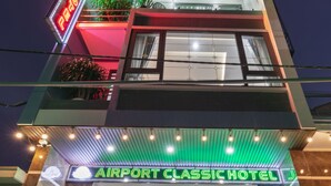 Exterior del Hotel Airport Classic con un letrero de neón, un mostrador de recepción y plantas en macetas.