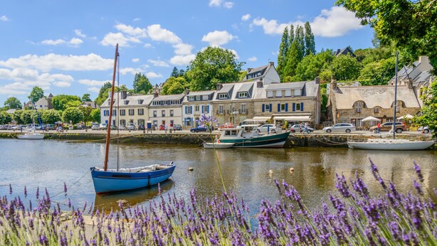 A waterfront scene with a row of traditional buildings, a blue boat docked near the shore, and lavender fields in the foreground. - Ty Kergui - Villa 400m from Tahiti sand beach (Névez)